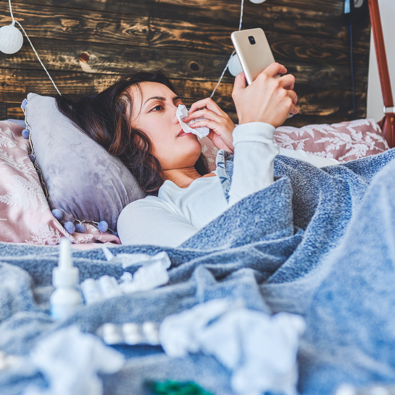 Frau liegt krank mit Schnupfen im Bett und betätigt ihr Smart Phone
