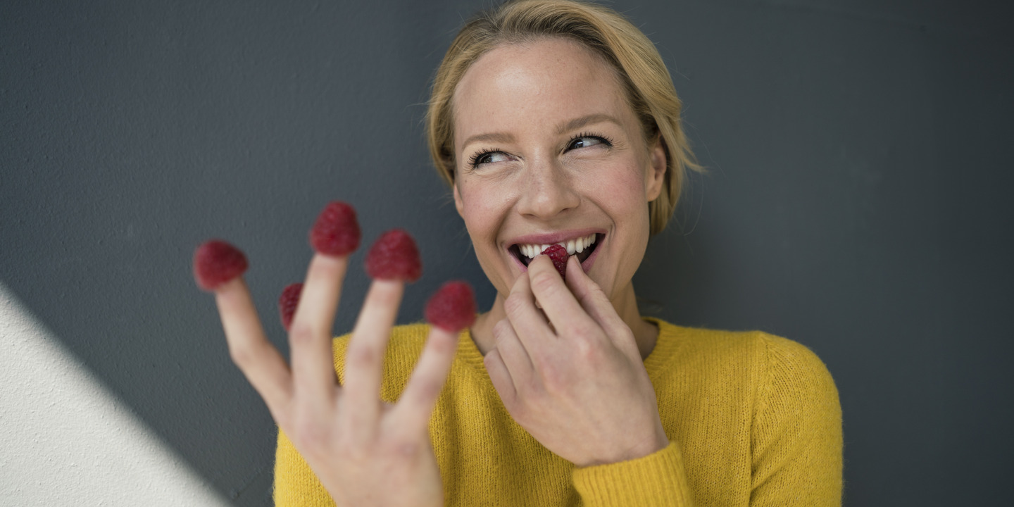 Lachende Frau isst Himbeeren, die auf ihren Fingerkuppen stecken