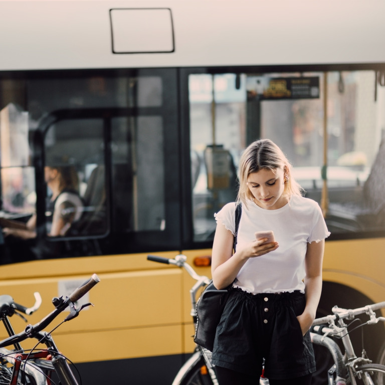 Junge Frau steht auf ihr Smartphone schauend vor einem gelben Bus