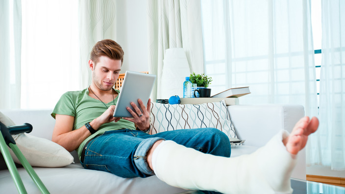 junger Mann sitzt mit einem eingegipsten Bein auf dem Sofa und bedient ein Tablet