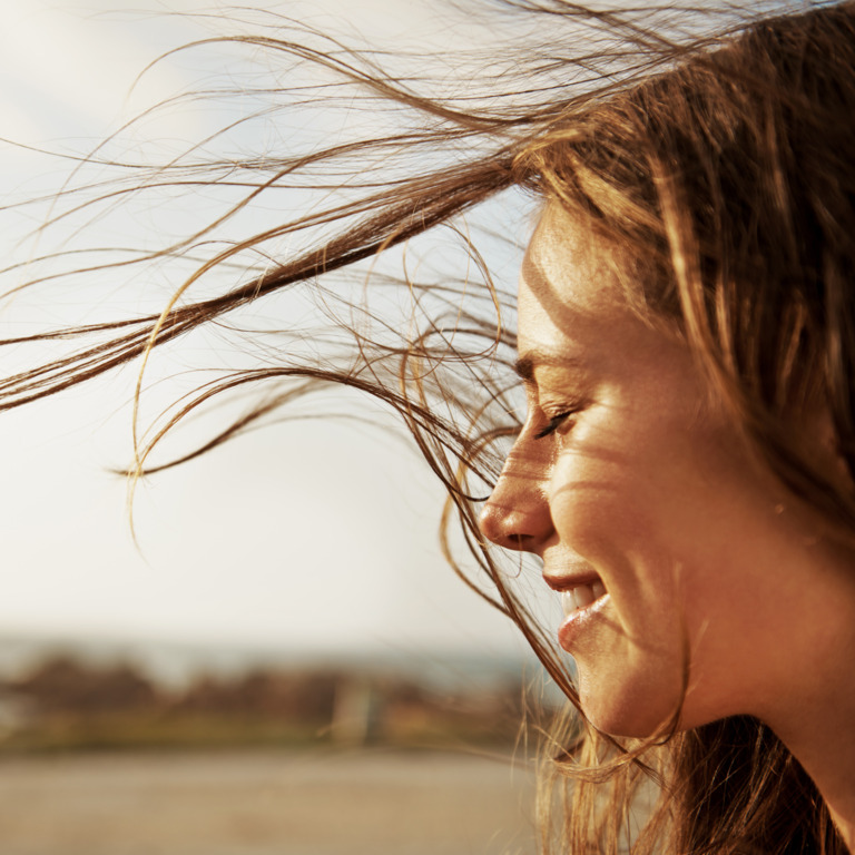 Frau steht auf offenem Feld und genießt, wie ihr der Wind durch die Haare fährt.