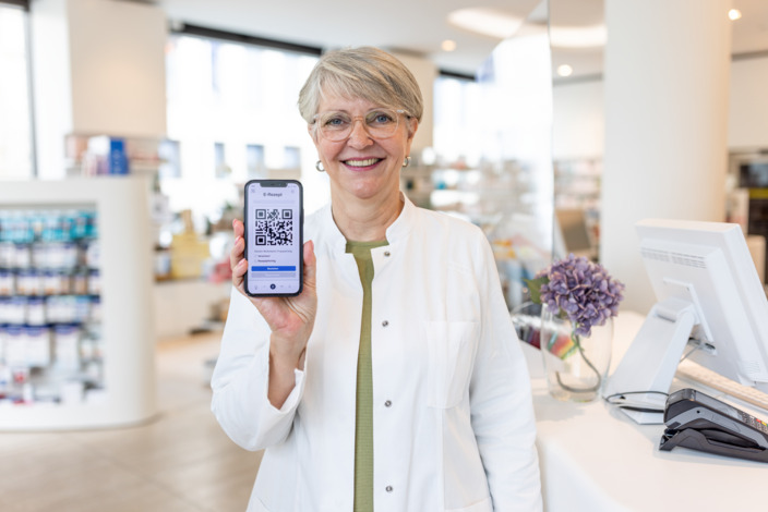 Neuerungen im Gesundheitswesen: Eine Pharmazeutin im weißen Kittel zeigt ein Handy, auf dem ein E-Rezept geöffnet ist.