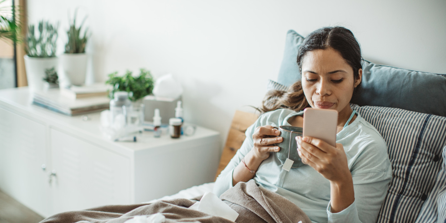 Eine Frau liegt  krank im Bett und hält in ihrer Hand ein Smartphone.