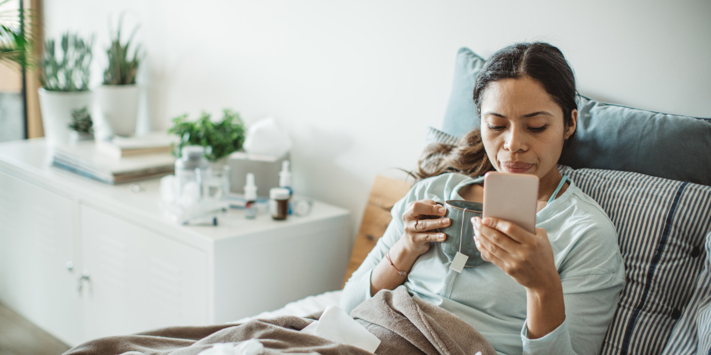 Eine Frau liegt krank im Bett und hält in ihrer Hand ein Smartphone.