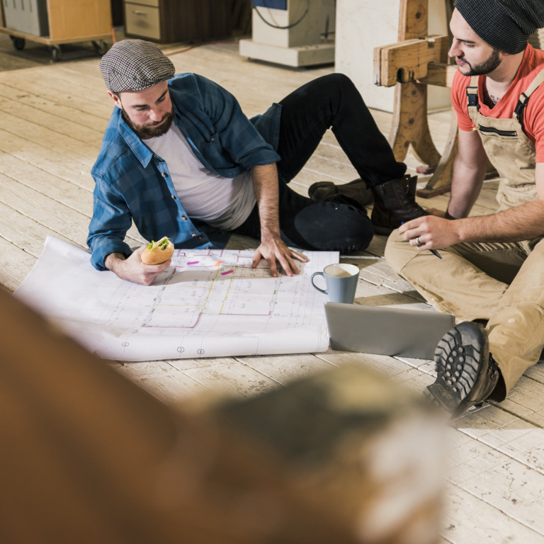 Männer in Arbeitskleidung hocken auf dem Fußboden und schauen gemeinsam auf einen Bauplan