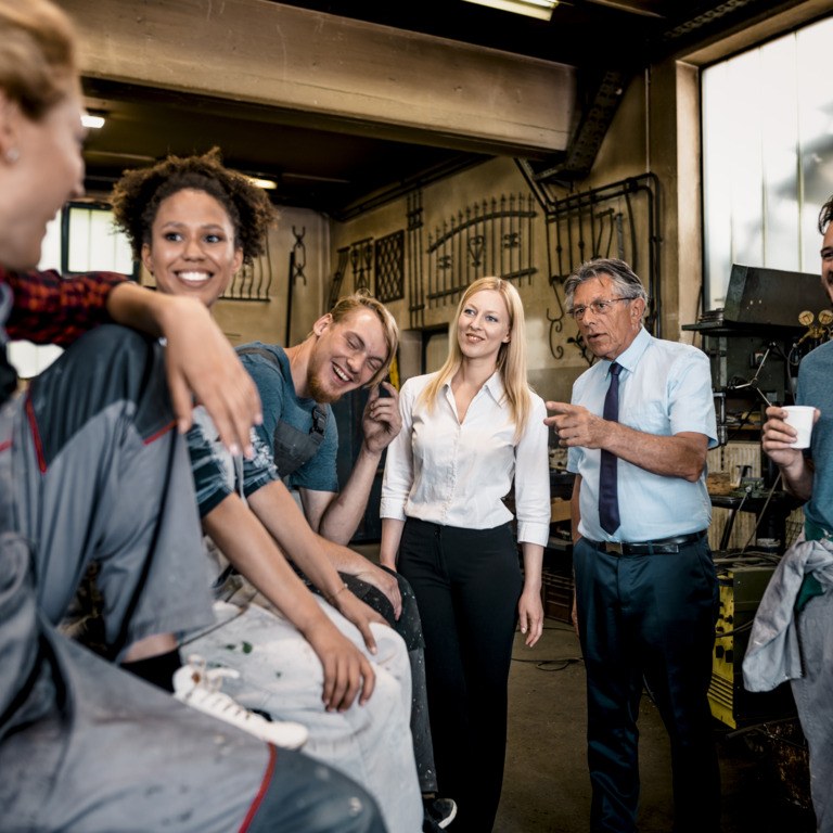 Eine Gruppe aus Frauen und Männern in Arbeitskleidung steht in einem Arbeitsraum. Ein Mann in Hemd und Krawatte zeigt auf einen der Männer, der Lacht.