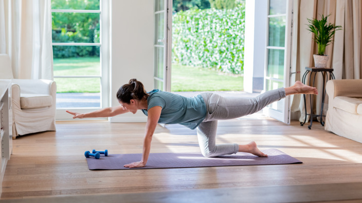 Frau kniet in ihrem Wohnzimmer auf einer Yogamatte und macht eine Yogaübung