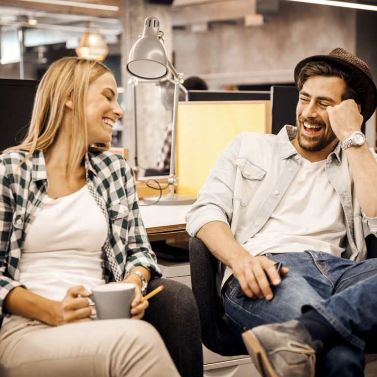 Junges Pärchen lacht am Arbeitsplatz miteinander.