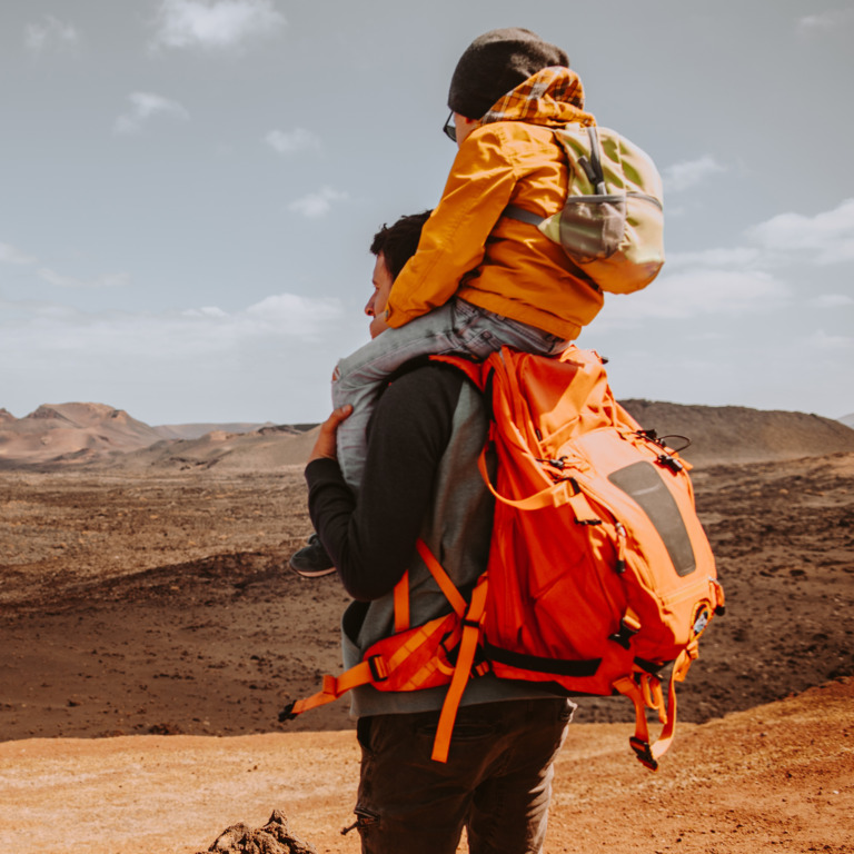Vater im Trekking-Outfit trägt seinen Sohn auf den Schultern und blickt in eine Wüstenlandschaft