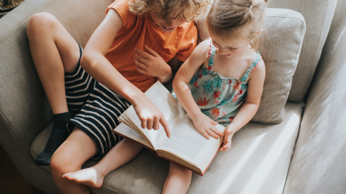 Zwei Kinder sitzen ein Buch lesend auf dem Sofa