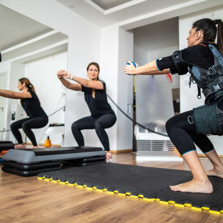 Drei Frauen, die EMS-Training durchführen und dabei Kniebeugen machen. 