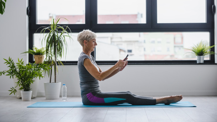 Frau schaut in ihr Smartphone am Boden auf einer Yogamatte sitzend