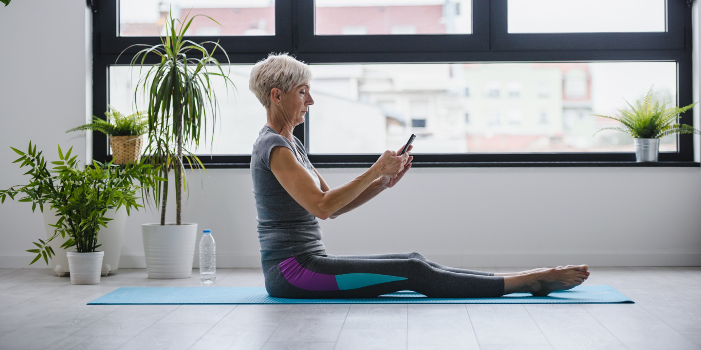 Frau hält ein Handy in der Hand und sitzt in Sportkleidung auf einer Yogamatte