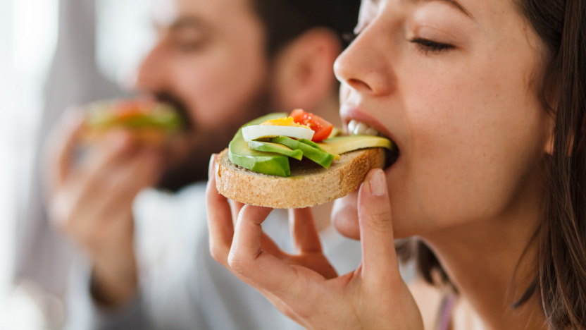 Ein junges Paar (Frau im Vordergrund) essen genüsslich ein Brot mit Avocado, Ei und Cherry-Tomaten belegt.