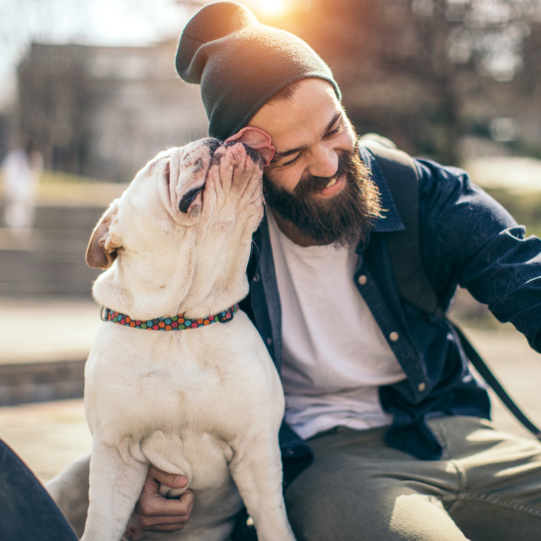 Ein junger Mann sitzt neben einem Hund in einem Park. Der Mann lacht, während ihm vom Hund das Gesicht geleckt wird und er ein Selfie macht.