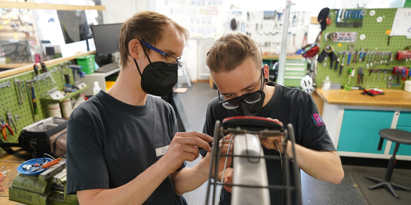 Vielfalt in der Fahrradwerkstatt: Zwei junge Erwachsene mit unterschiedlichem Hintergrund reparieren gemeinsam ein Fahrrad.