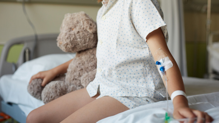 Mädchen sitzt auf dem Rand eines Krankenhausbettes und hält ihren Teddy im Arm