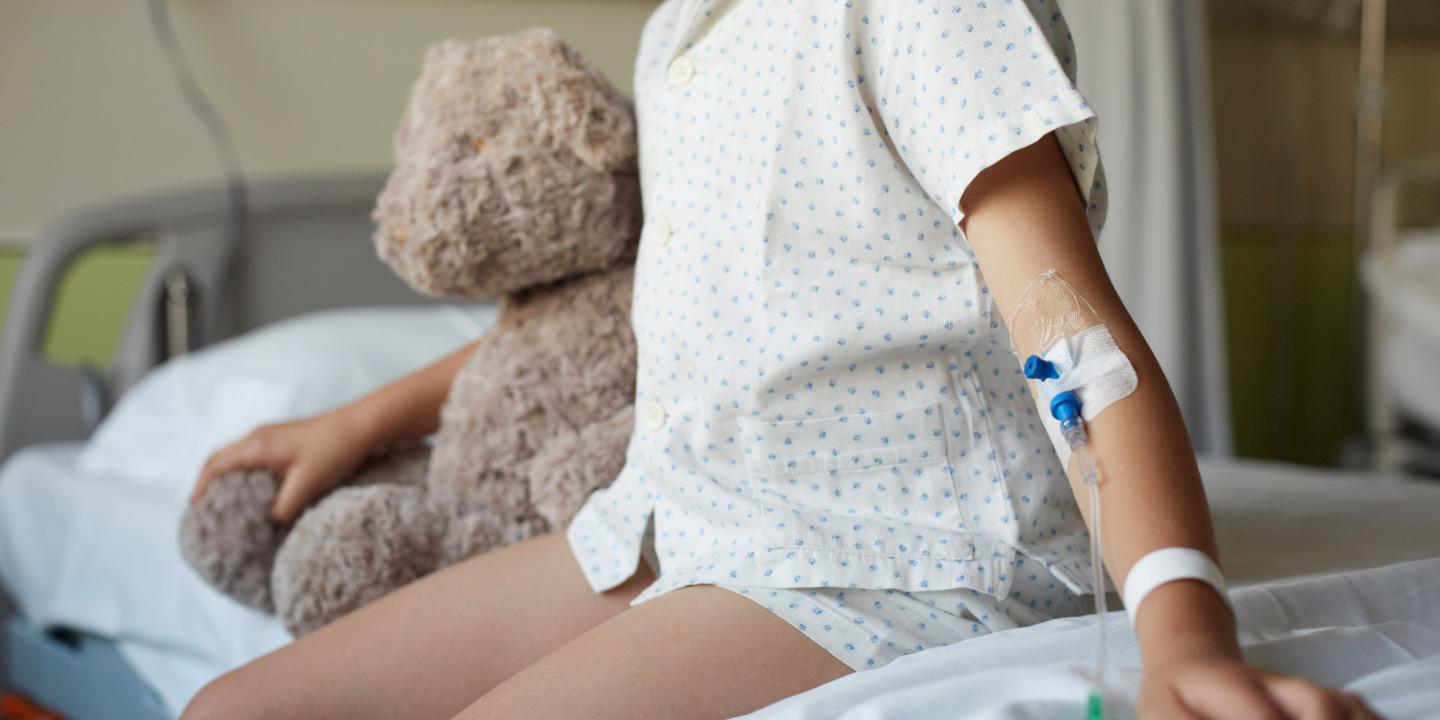 Kind sitzt in Palliativstation auf dem Bett mit Teddy im Arm.