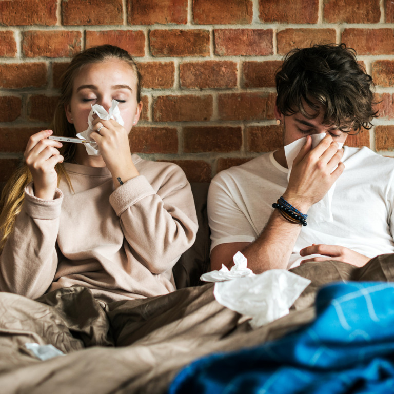 Frau und Mann liegen krank im Bett und schnäuzen in Taschentücher.