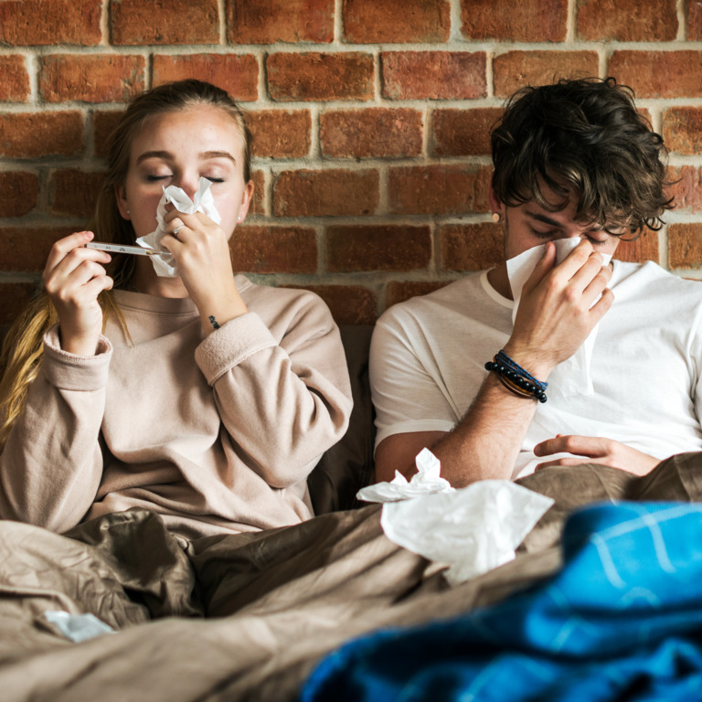 Frau und Mann sitzen im Bett und putzen sich die Nase.