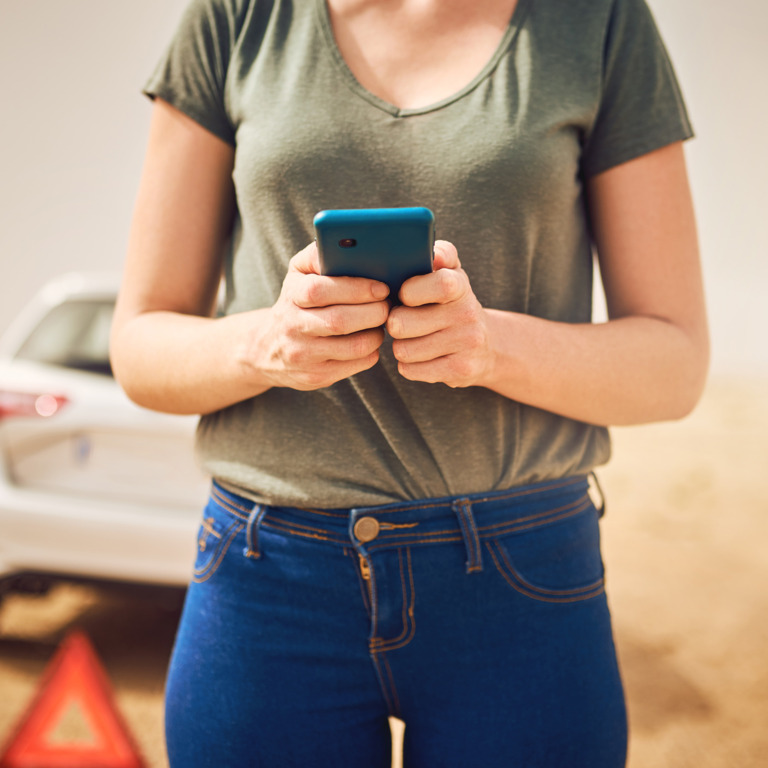 Frau steht mit einer Auto-Panne am Straßenrand und ruft Hilfe mit dem Smartphone.