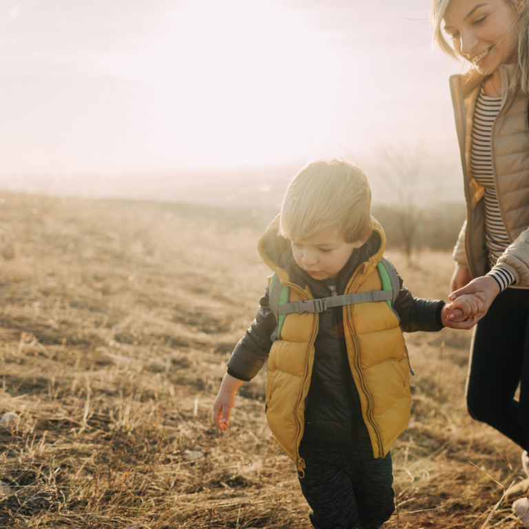 Eine Familie geht wandern, im Hintergrund trägt eine Person ein Kind auf den Schultern, im Vordergrund führt eine Frau ein Kind an der Hand.