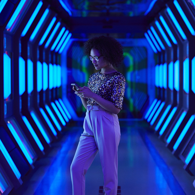 Junge Auszubildende steht in einem neonblau-beleuchtetem Gang und tippt auf ihrem Handy