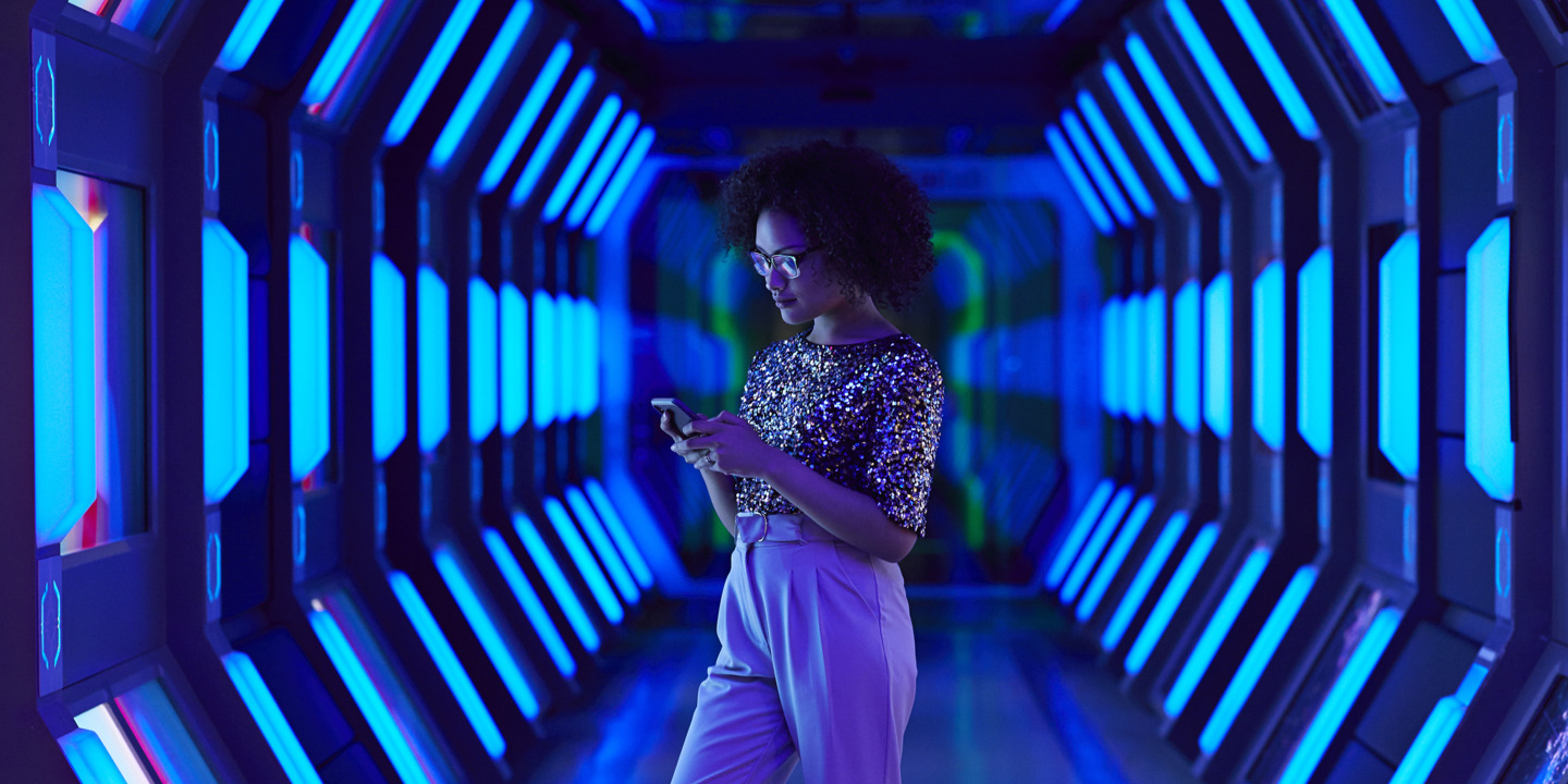 Junge Auszubildene steht in einem neonblau-beleuchtetem Gang und tippt auf ihrem Handy