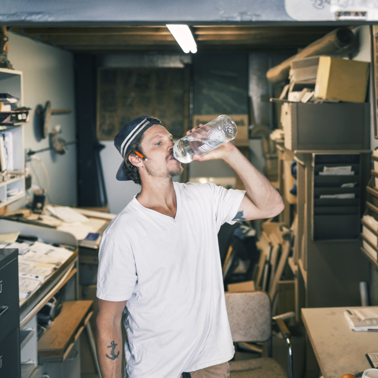 Junger Mann, der in einer Werkstatt aus einer Wasserflasche trinkt.