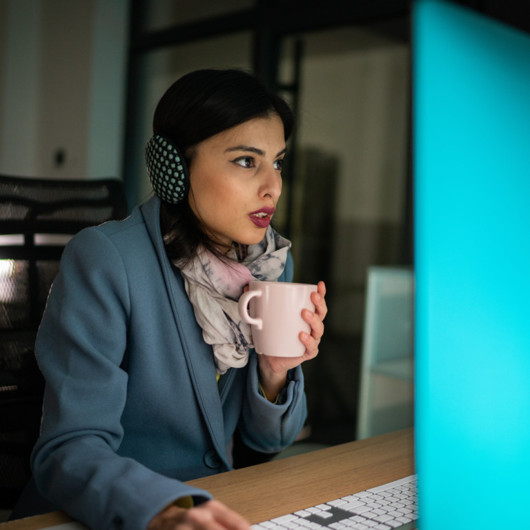 Mitarbeiterin am Computer mit Ohrwärmern auf dem Kopf und Teetasse in der Hand