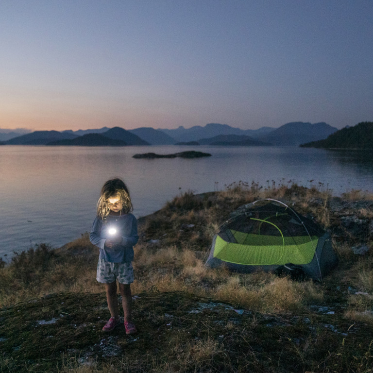kleiner Junge steht in der Abenddämmerung am Ufer eines Sees und leuchtet mit seiner Taschenlampe