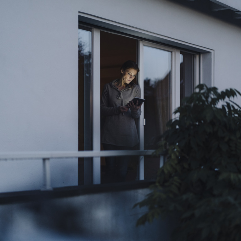Frau steht an offenem Fenster und liest auf ihrem Tablet