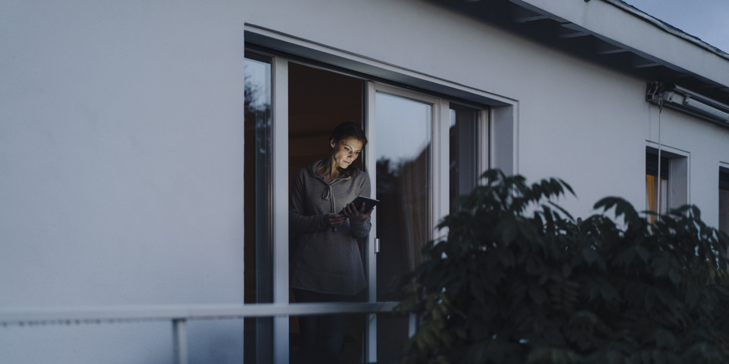 Frau steht abends an geöffnetem Fenster und liest auf ihrem Tablet