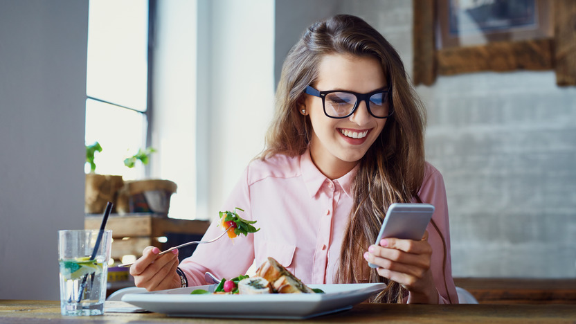 junge Frau isst etwas und schaut dabei auf ihr Smartphone