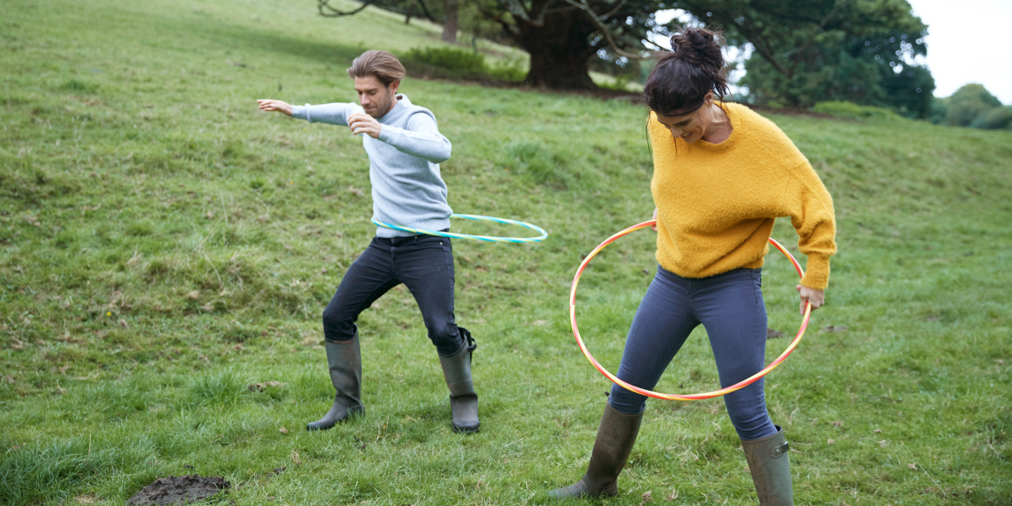 Mann und Frau in Gummistiefeln machen Hoola Hoop Übungen auf einer saftig grünen Bergwiese
