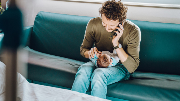 Mann mit Baby sitzt auf türkiser Couch und telefoniert