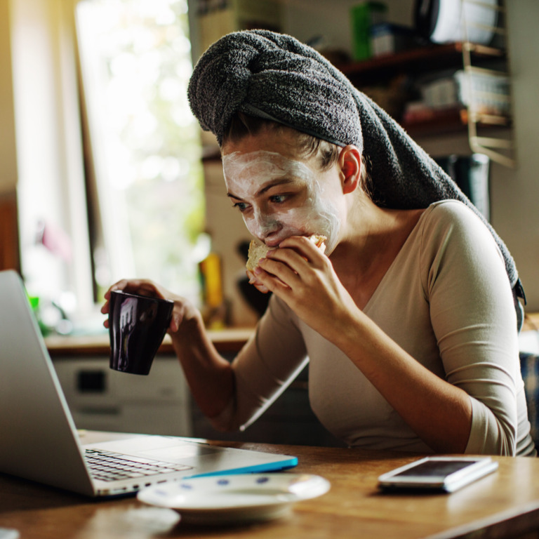 Eine Frau arbeitet mit Gesichtsmaske und Handtuch im Haar am Laptop, während sie gleichzeitig ein Sandwich isst und Kaffee trinkt.