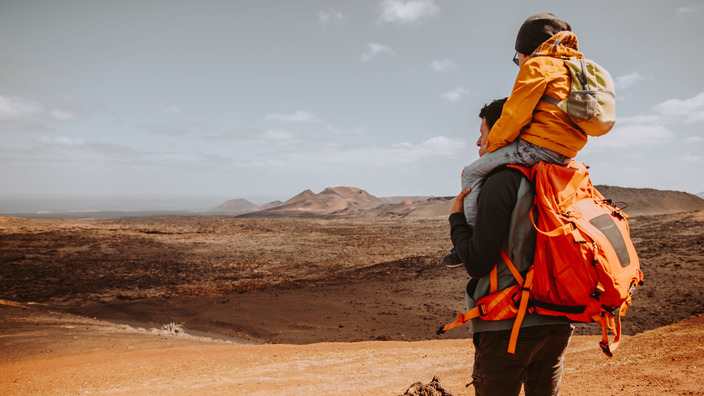 Vater im Trekking-Outfit trägt seinen Sohn auf den Schultern und blickt in eine Wüstenlandschaft