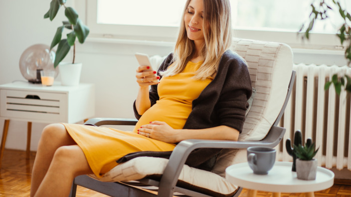 Schwangere sitzt in einem Sessel und nutzt die BabyCare-App auf ihrem Smartphone
