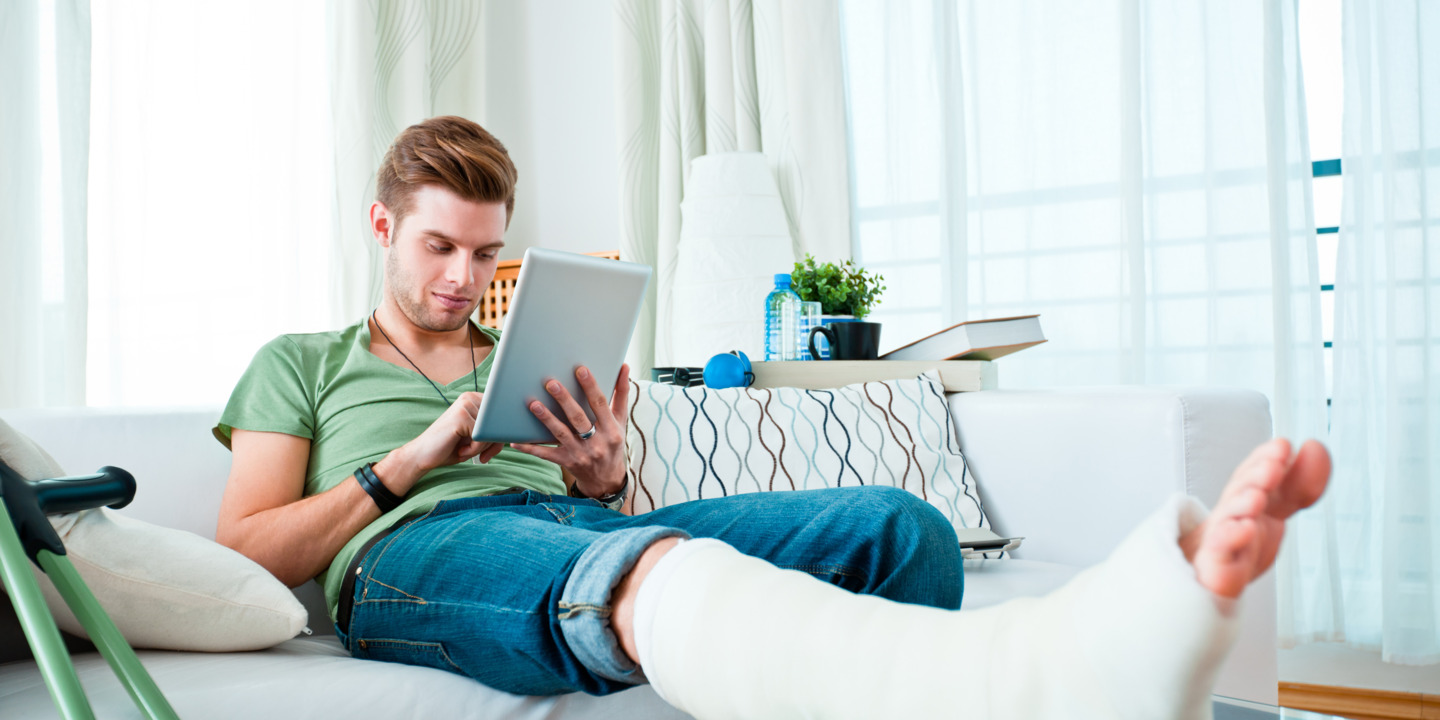 junger Mann sitzt mit einem eingegipsten Bein auf der Couch und bedient ein Tablet