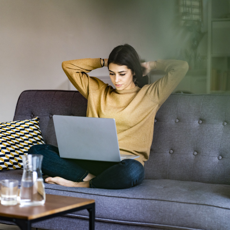 Junge Frau sitz mit dem Laptop auf dem Schoß auf der Couch