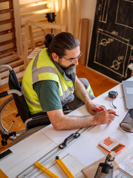 Mann im Rollstuhl arbeitet an Bauzeichnungen am Schreibtisch.