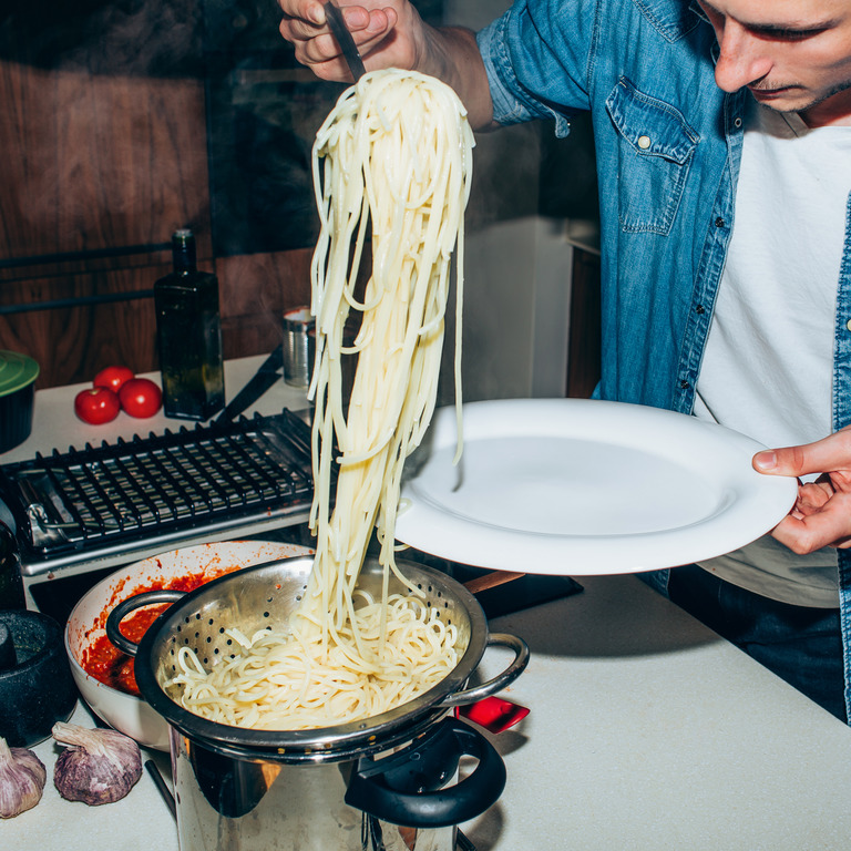 Junger Mann im Jeanshemd füllt sich Spaghetti aus dem Topf auf den Teller