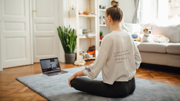 Junge Frau, die vor einem Laptop auf dem Teppich eine Yoga-Übung macht.