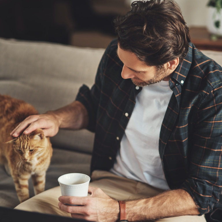 Mann im Karohemd sitzt mit einer Tasse auf der Couch und streichelt eine rote Katze