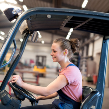 Junge Frau in Blaumann und rotem Shirt fährt einen Gabelstapler.