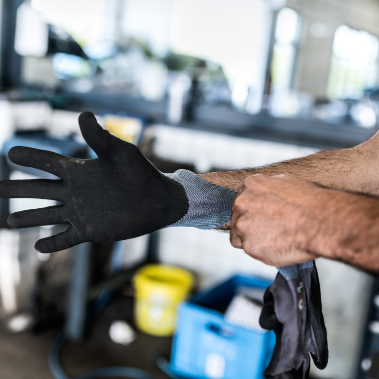 Arme eines Mannes, der sich in einer Werkhalle schwarze Handschuhe anzieht.