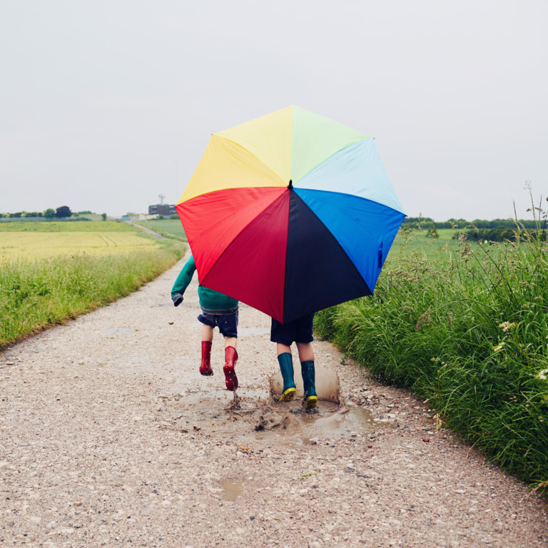 zwei Kinder in Gummistiefeln laufen mit einem großen regenbogenfarbenen Schirm über einen Feldweg im Regen