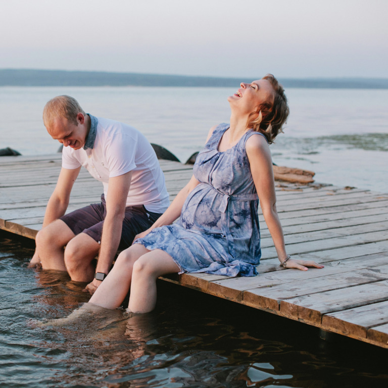 Eine schwangere Frau und ein Mann sitzen auf einem Steg und lassen die Beine im Wasser baumeln