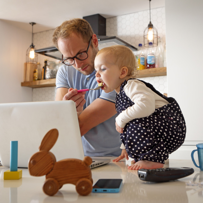 Ein Mann füttert ein kleines Kind. Dabei schauen der Mann und das Kind auf den Bildschirm eines Laptop.